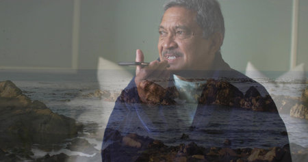 Holding senior man speaking into smartphone at home, in dark jacket, with cushion, rocks and waves....
