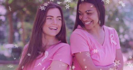 Smiling two women wearing pink vneck tees posing in park, showing ribbon pins and snowflakes. Outdoor, duo, friendship, awareness, fundraising, campaign, natureの写真素材