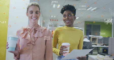 Walking two women in peach-blouse and mustard-top in office holding coffee cups, tech overlay. Collaboration, workspace, modern, digital, interface, hexagon, beverageの写真素材