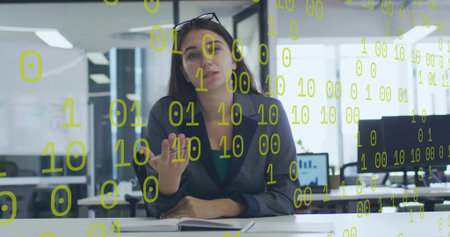 Presenting woman in dark blazer, teal top gesturing at desk in office, notebook, binary overlay. Data, digits, visualization, analytics, business, workspace, monitorsの写真素材