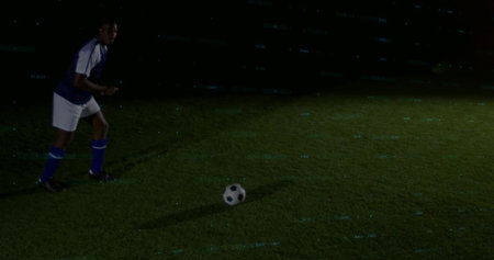 Preparing soccer player in blue kit and cleats, eyeing ball on pitch under floodlights, copy space. Footballer, night, stadium, turf, shadows, spotlight, jerseyの写真素材