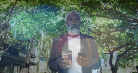 Standing midadult man wearing blazer white shirt on tree-lined walk, checking phone, holding coffee. Professional, urban, jacket, bag, smartphone, earphones, takeawayの写真素材