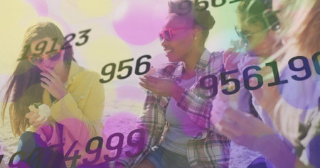 Laughing four women in yellow jacket plaid shirt shades talking on sandy beach, with number bokeh....