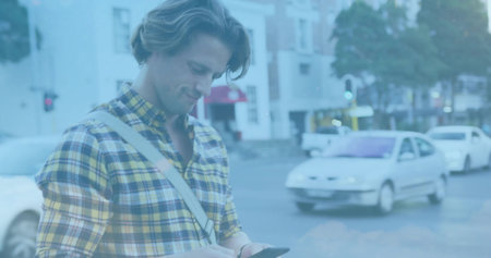 Standing man wearing yellow-plaid shirt, looking at phone at city corner, with crossbody bag, cars....