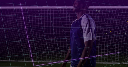 Standing soccer player wearing blue bib, white tee and shorts on turf by goal, purple grid. Athlete,...
