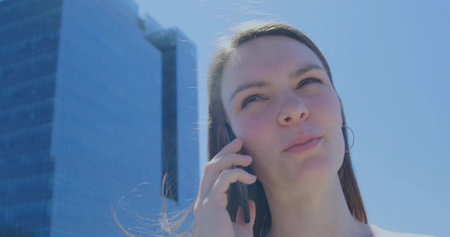 Talking woman on smartphone in downtown near glass towers light top hoop earring sunlight on hair. Urban, professional, portrait, phone, backlit, reflective, skyscraperの写真素材
