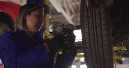 Operating female mechanic in blue coveralls and black gloves using impact wrench on wheel at garage....