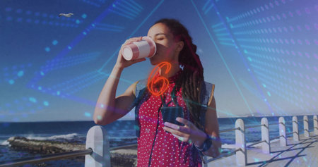 Sipping woman red-polka-top denim-vest holding paper and travel cups on promenade. Coastal, boardwalk, backpack, denim, casual, sunny, sereneの写真素材