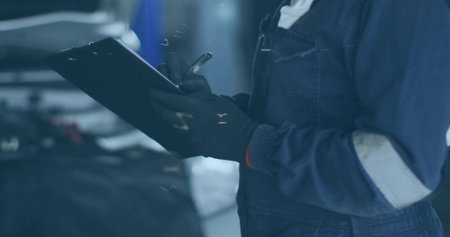 Checking woman mechanic writing on clipboard with pen in garage, open hood, coveralls, gloves. Technician, automotive, workshop, engine, inspection, reflective, industrialの写真素材