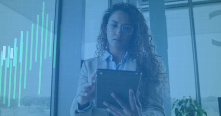 Standing woman checking tablet by large windows with teal chart overlay, in blazer and glasses....