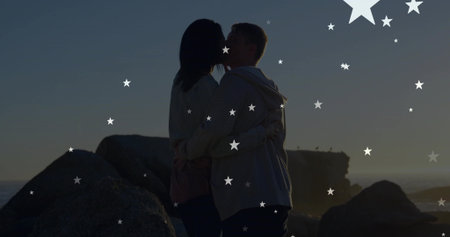 Kissing couple embracing on rocky shore at dusk, in hoodies, boulders, cliff, horizon, stars. Romance, silhouette, sunset, coastal, seascape, twilight, moodyの写真素材