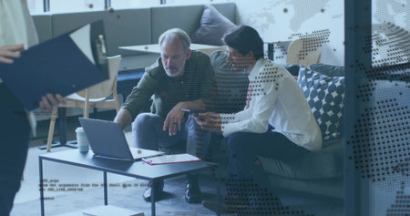Discussing two seated men in white shirt pointing at laptop on lounge sofa, with clipboard....