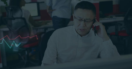 Holding phone, man wearing light shirt looking at monitor at desk with red chairs, chart. Office,...