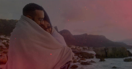 Embracing couple sharing light blanket on rocky shoreline at dusk, man wearing eyeglasses. Coastal,...