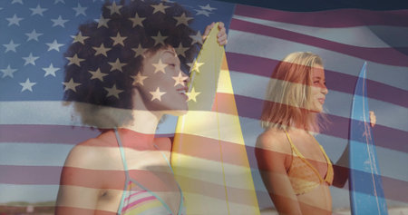 Holding yellow and blue surfboards, two women in bikinis standing on beach, American flag overlay. Duo, surfers, boards, swimwear, coast, shoreline, oceanの写真素材