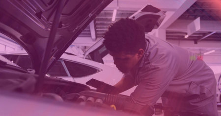 Leaning mechanic in gray work shirt inspecting engine bay under raised hood at shop, holding phone. Garage, technician, workshop, tools, diagnostic, purple, lightingの写真素材