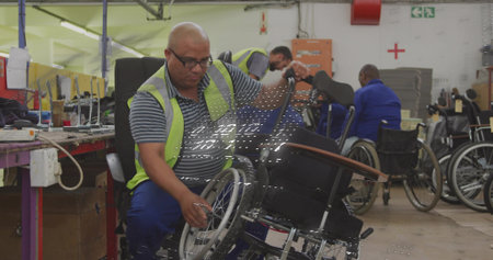 Working technician adjusting wheelchair wheel at workshop, wearing hi-vis vest and striped polo. Mobility, assembly, factory, industrial, tools, safety, teamworkの写真素材