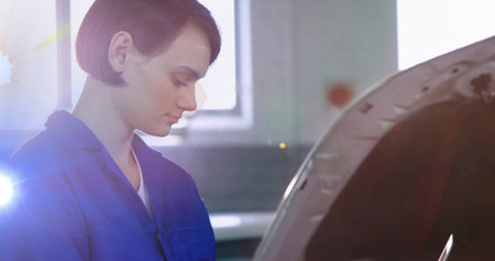 Inspecting woman technician in blue coveralls leaning over car with open hood in garage, lens flare....