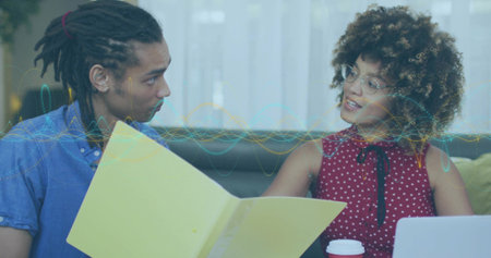 Discussing peers holding yellow folder on sofa in blue shirt red top with laptop, waveform, glasses. Collaboration, teamwork, lounge, casual, documents, computer, daylightの写真素材