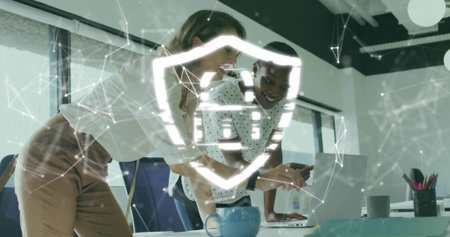 Leaning two business women pointing at silver laptop in office, holographic shield and blue mug....
