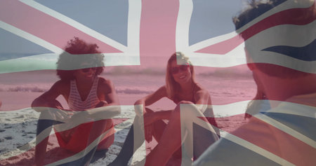 Sitting five adults socializing on Union Jack-tinted beach by waterline, wearing swimwear. Friends, casual, seaside, flag, lifestyle, leisure, socialの写真素材