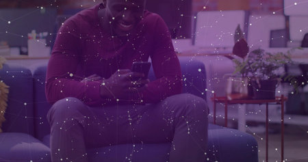 Sitting African American man wearing maroon shirt and watch, checking smartphone on sofa in lounge....