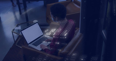 Typing woman wearing maroon top and face mask using laptop in brown lounge armchair, dotted lights....