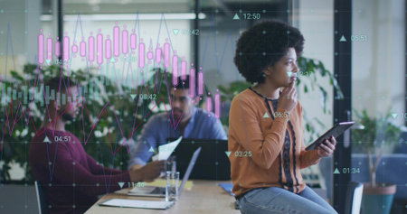Holding tablet, woman orange top sitting on right table edge office using laptop charts, copy space. Collaboration, teamwork, analytics, data, strategy, focus, daylightの写真素材