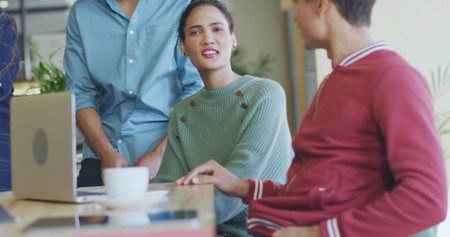 Speaking woman in mint-green sweater engaging colleagues at bright workspace, laptop and coffee cup....