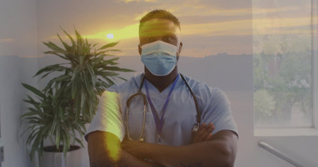 Standing clinician wearing blue scrubs and surgical mask in clinic, with stethoscope, blue lanyard. Healthcare, professional, medical, sunset, glow, ambient, plantの写真素材