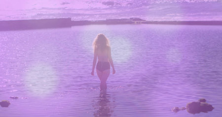 Walking woman in swim bottoms wading into shallow water toward horizon, with rocks and lens flare. Silhouette, stroll, coastal, seascape, beach, magenta, violetの写真素材