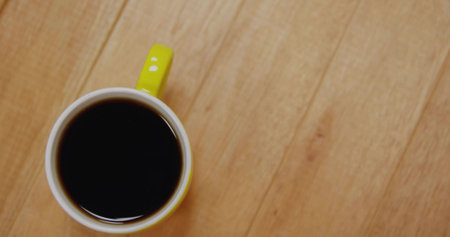 Sitting yellow-handled ceramic mug showing coffee glint on light wood tabletop, copy space. Topdown, flatlay, cup, beverage, cafe, kitchen, warmの写真素材