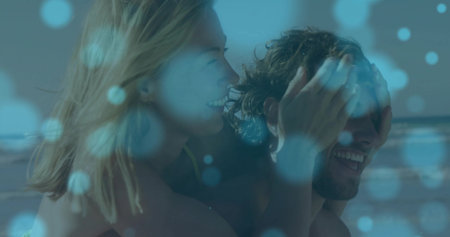 Giving woman covering man's eyes carrying him at beach shoreline, swimsuit top, trunks, blue bokeh....