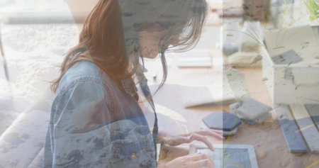 Leaning forward, mid-adult woman tapping tablet on wooden desk, wearing denim jacket, ocean overlay. Remote, study, homeoffice, workspace, sunlit, serene, doubleexposureの写真素材