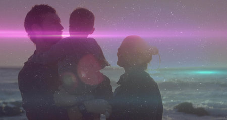 Holding baby, parents facing on rocky shore at dusk, wearing jackets, magenta lens flare....