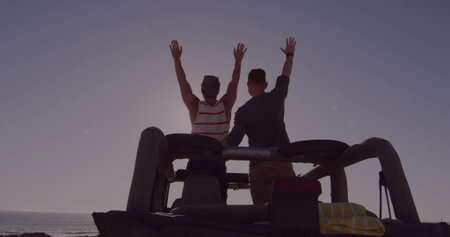 Raising arms, two men standing on open-top jeep rear at ocean dusk, wearing striped tank, towel. Silhouette, sunset, coastal, backlit, sea, horizon, travelの写真素材