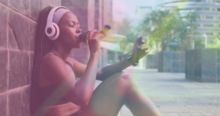 Drinking woman in sports bra and shorts, sitting on paved walkway with bottle and smartphone. Outdoor, urban, fitness, hydration, headphones, watch, phoneの写真素材