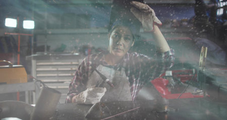 Welding woman wearing plaid top, protective apron and heavy gloves handling torch at workshop bench. Metalworker, fabrication, industrial, tools, lighting, sparks, focusの写真素材