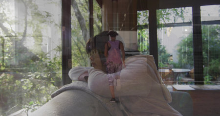 Reclining woman in sweater on couch at home, mirroring walking child wearing pink pack in glass. Family, parent, caregiver, garden, outdoor, daylight, reflectionの写真素材