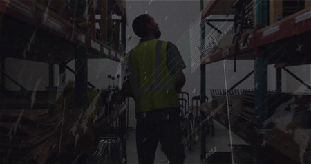 Standing warehouse worker wearing hi-vis vest checking shelves in dimly lit aisle with pallets. Industrial, storage, inventory, safety, gritty, lowlight, concreteの写真素材