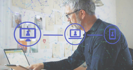 Working senior man wearing glasses, dark sweater using laptop, tablet in home office, network icons....