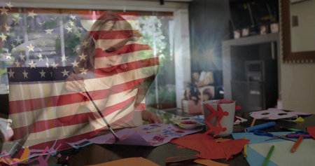 Painting child in light-colored top using paintbrush at home craft table, flag overlay, paints. Girl, art, colors, daylight, window, garden, livingroomの写真素材