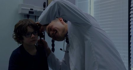 Leaning Asian doctor in lab coat examining child with otoscope on exam chair in clinic stethoscope. Pediatric, examination, physician, otoscopy, ear, equipment, lightingの写真素材