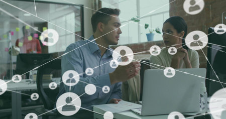 Discussing man in blue shirt, woman in sweater examining laptop at desk in office, network overlay. Collaboration, teamwork, meeting, partition, daylight, brick, computerの写真素材