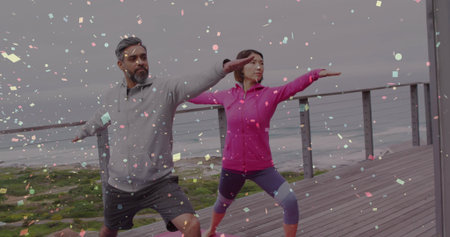Doing pair holding Warrior pose on wooden deck over ocean, wearing bright sportswear with yoga mats....