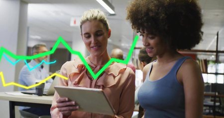 Reviewing tablet, two women wearing peach blouse and blue top in open office with line graphs. Colleagues, collaboration, data, charts, analytics, workspace, modernの写真素材