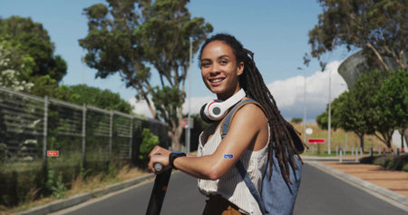 Smiling woman holding electric scooter on campus road, wearing striped top backpack headphones. Female, emobility, bag, headset, suburban, chainlink, treesの写真素材