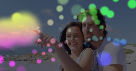 Pointing sleeveless woman and tee-clad man holding, gazing at horizon on rocky shore, with bokeh. Couple, beach, coastline, daylight, sunshine, romance, affectionの写真素材