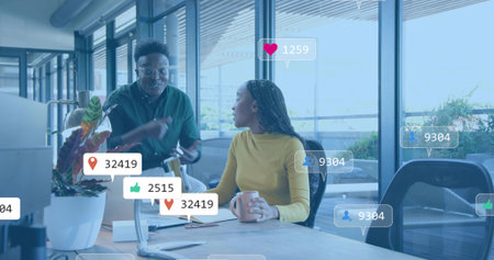 Gesturing pair in green-shirt and mustard-top talking at office desk with laptop mug social-icons. Collaboration, teamwork, conversation, brainstorm, digital, modern, brightの写真素材