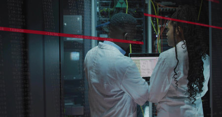 Two techs in white lab coats fixing servers in server aisle with yellow cables and laptop....